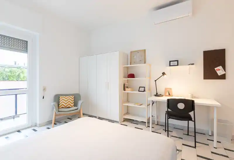 Bright, modern bedroom with a comfortable bed in the foreground, a wardrobe and shelving, and a simple desk by the wall — ideal for a student or professional seeking a tidy, well-lit room.