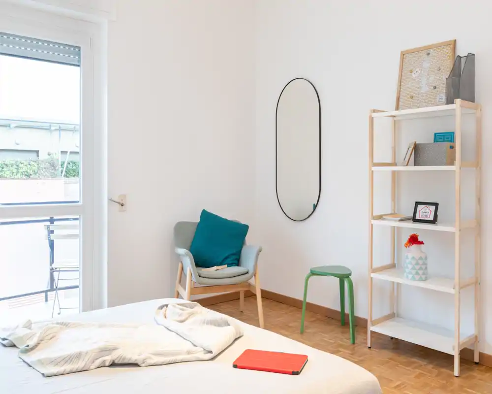 Camera da letto luminosa e minimale con letto in primo piano, una poltrona accogliente con cuscino turchese, specchio a parete e scaffalatura: ideale per un annuncio che valorizzi la luce naturale e un arredamento semplice.