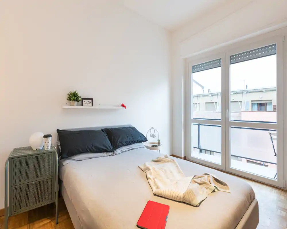 Camera da letto luminosa e minimalista con letto matrimoniale, biancheria ordinata e ampia finestra che lascia entrare molta luce naturale. Un piccolo comodino e una mensola completano l'aspetto moderno e tranquillo, perfetto per annunci di affitto.