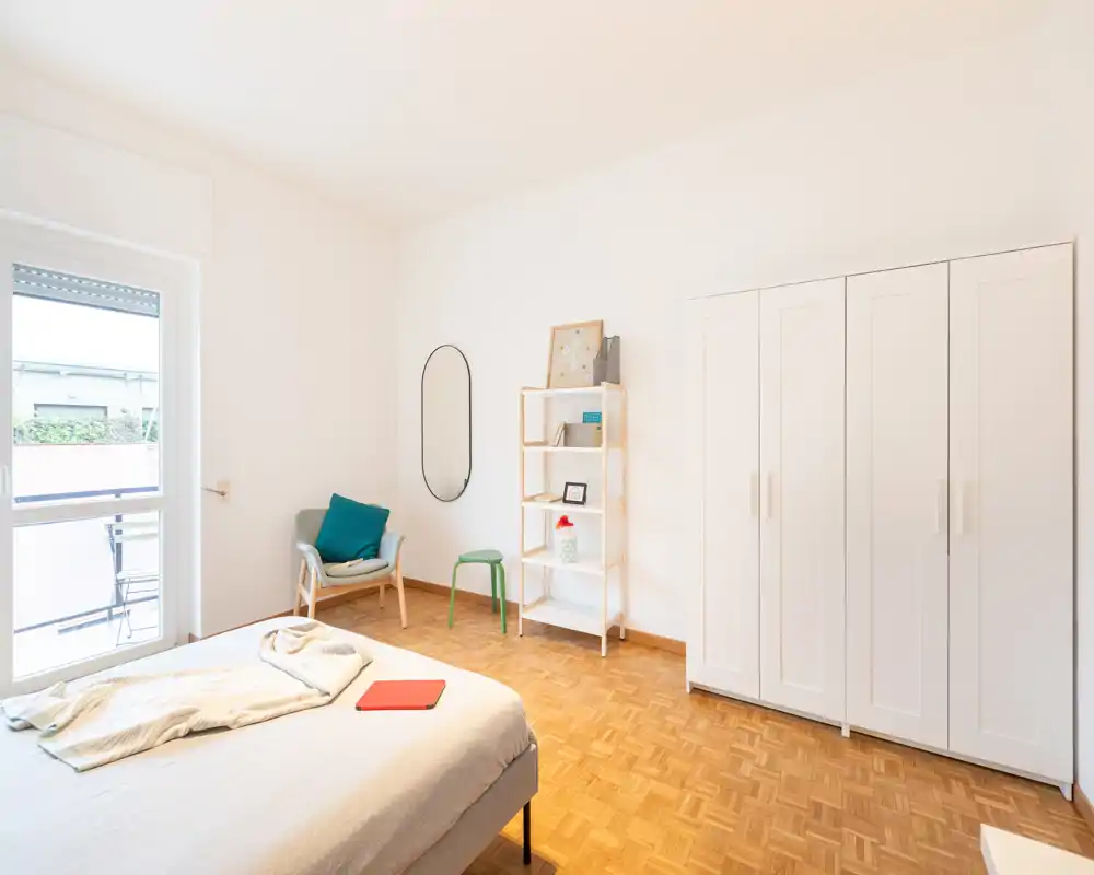 Camera da letto luminosa e minimalista con letto ben sistemato, armadio bianco, scaffalatura e una comoda poltrona vicino alla finestra con luce naturale.