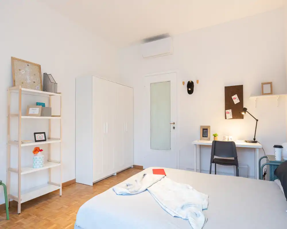 Camera da letto luminosa e minimal con un ampio letto in primo piano, un armadio bianco, mensole e una piccola scrivania con lampada — ambiente moderno e accogliente.