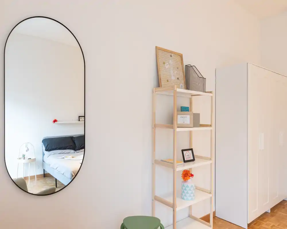 Angolo di camera da letto luminoso e minimale con letto matrimoniale visibile nello specchio ovale, mensole semplici e un armadio bianco — ideale per un annuncio moderno.
