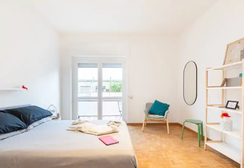 Camera da letto luminosa e minimalista con un ampio letto in primo piano, luce naturale dalla finestra/portafinestra, una poltrona accogliente e scaffalatura aperta—perfetta per un annuncio moderno.
