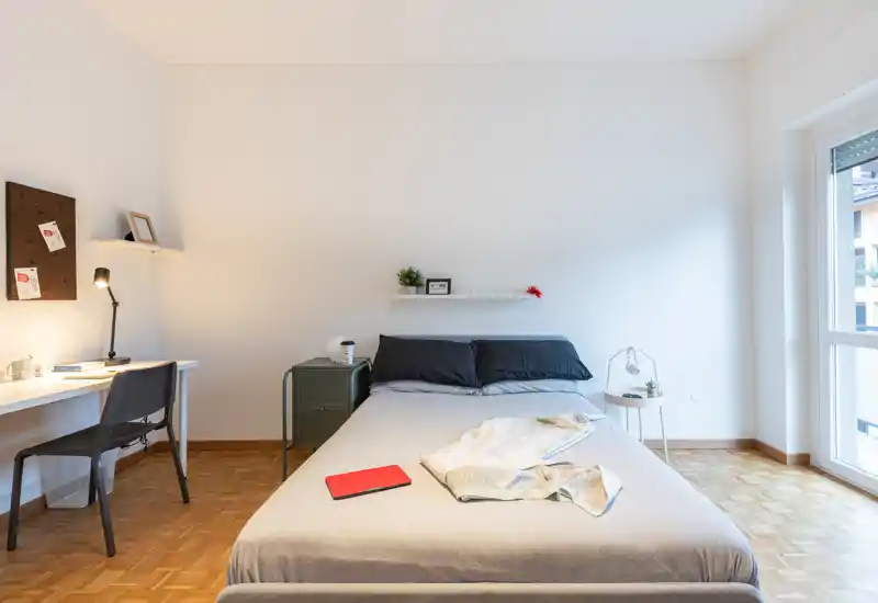 Camera da letto luminosa e minimalista con un ampio letto al centro dell’inquadratura, comodini, una scrivania sulla sinistra e una grande finestra che illumina l’ambiente. Toni neutri e arredi semplici creano un’atmosfera calma e ariosa ideale per annunci.