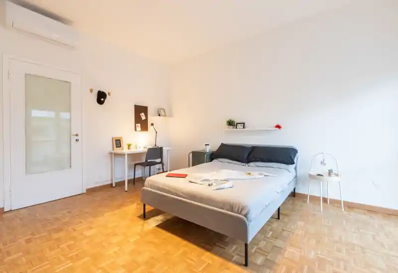 Camera da letto luminosa e minimal con letto matrimoniale, comodini e una piccola scrivania nell'angolo. Pavimento in parquet e pareti neutre creano un ambiente tranquillo e accogliente, perfetto per annunci.