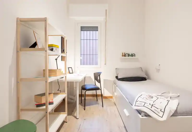Camera singola compatta con letto ordinato, una scrivania vicino alla finestra e scaffalatura aperta — luminosa, minimal e funzionale.