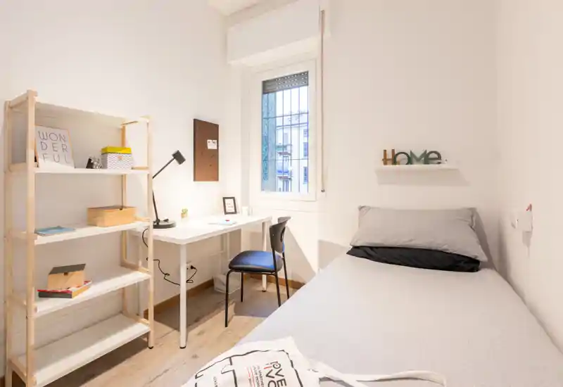 Camera compatta e luminosa con letto singolo, scrivania e scaffalatura—ideale per studenti o viaggiatori soli. Stile moderno pulito e luce naturale la rendono accogliente.