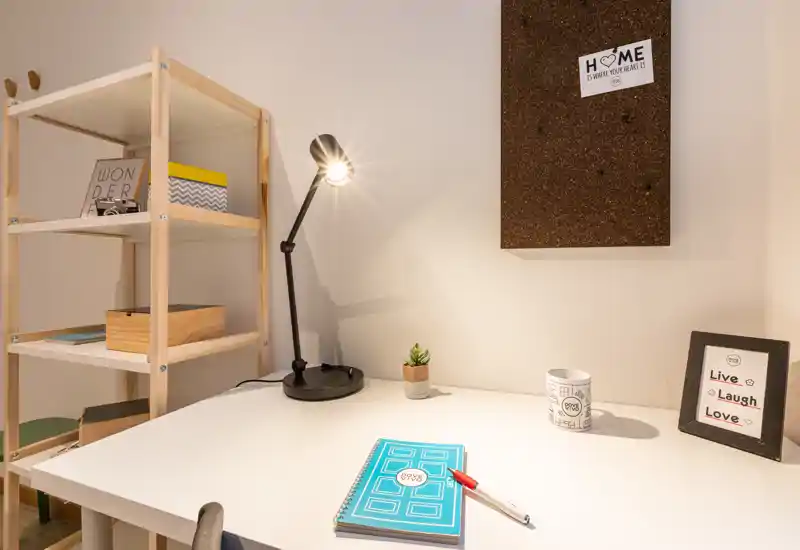 Bright, tidy workspace with a white desk, adjustable black desk lamp, shelving unit and small decorative items — ideal for studying or remote work.