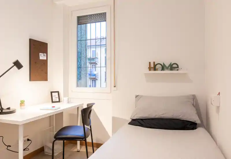 Camera singola compatta e luminosa con letto ordinato, mensola e scrivania vicino alla finestra — ideale per uno studente o single.