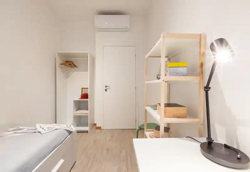 Camera da letto semplice e luminosa con letto singolo visibile, scaffalatura aperta e una lampada da scrivania — stile minimal ideale per affitti brevi.