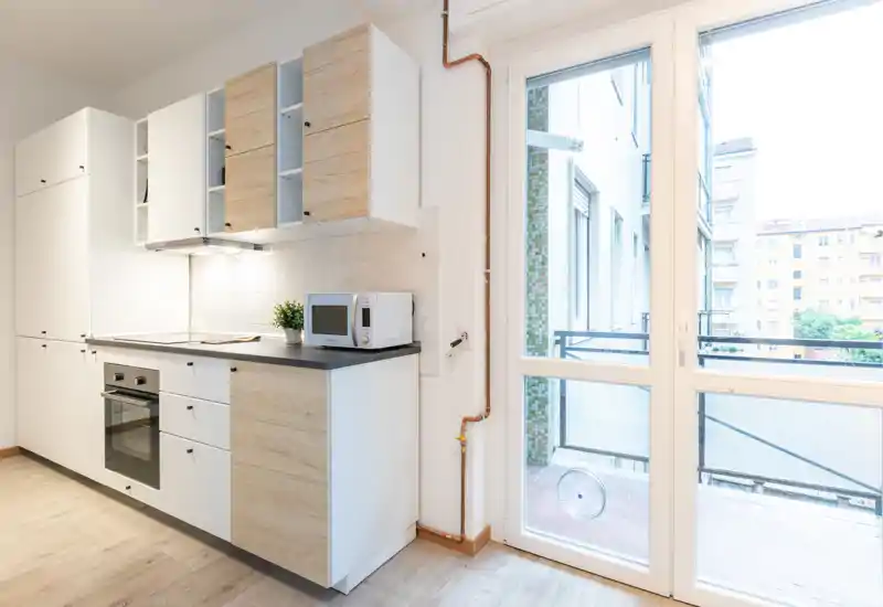 Bright, compact modern kitchen with white and light-wood cabinets, built-in oven and a microwave on the countertop. Large glass door opens to a small balcony, providing abundant natural light.