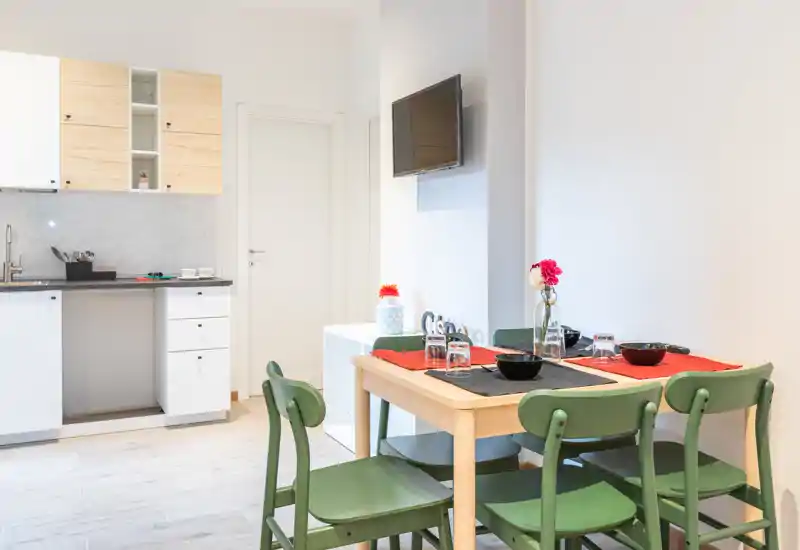 Bright, compact dining area next to a small kitchenette. A wooden table set for four with green chairs and simple tableware creates a cozy, functional space ideal for short stays.
