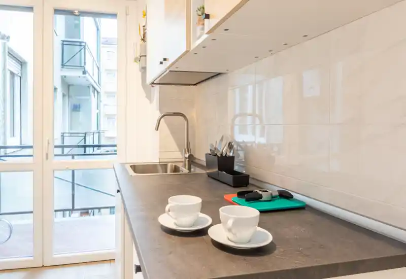Modern compact kitchen with a dark countertop, stainless sink and natural light from the glass door; two cups on saucers add a lived-in touch.