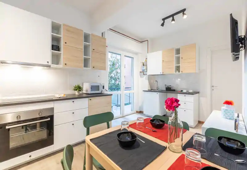 Bright, modern kitchen with built-in oven, white and wood cabinets, and a small dining table set for four. Large window/door brings natural light and a pleasant view to the space.