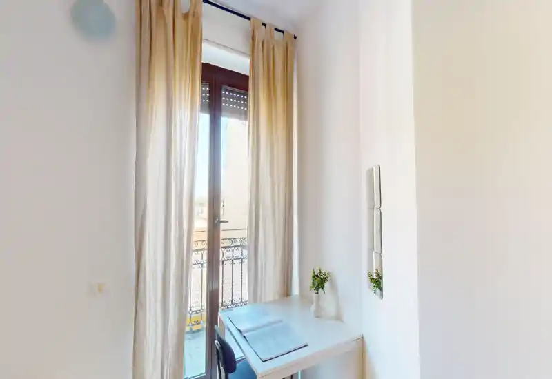 Angolo luminoso con scrivania davanti a una porta finestra che si apre sul balcone; tende leggere filtrano la luce naturale e due piantine danno un tocco fresco.