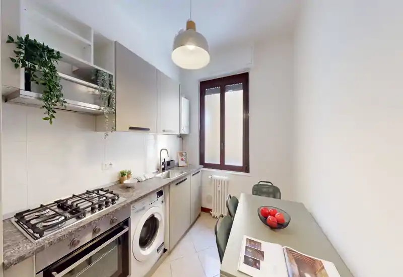 Cucina moderna compatta con piano a gas, forno e lavatrice sotto il bancone; un piccolo piano pranzo con sedie vicino alla finestra, luce naturale e finiture semplici e contemporanee.