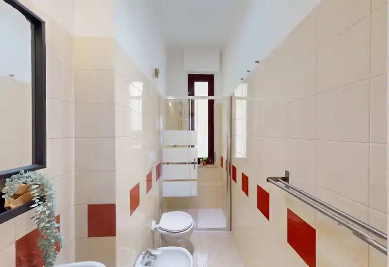 Bagno compatto e moderno con piastrelle a parete, bidet e WC, box doccia in vetro e una finestra verticale stretta che fornisce luce naturale.