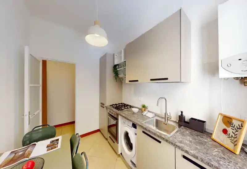 Cucina moderna compatta con piano lavoro effetto marmo, piano cottura a gas, forno incassato e lavatrice. Ambiente luminoso e ordinato con pensili e piccoli dettagli decorativi.