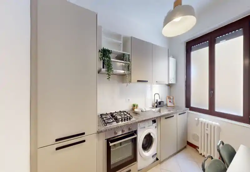 Cucina moderna compatta con elettrodomestici integrati, piano cottura a gas, forno e lavatrice; finestra luminosa e mobili neutri creano uno spazio pulito e funzionale.