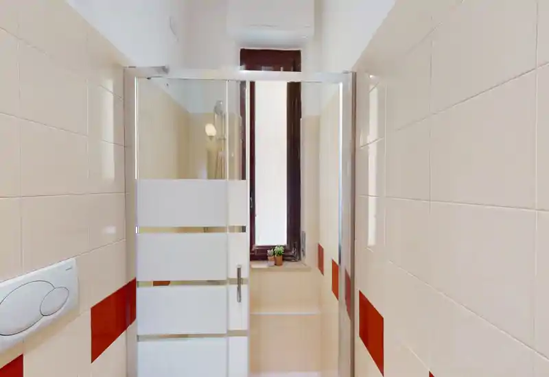 Bagno compatto e ben illuminato con box doccia in vetro, piastrelle bianche con dettagli rossi e una piccola finestra che fornisce luce naturale.