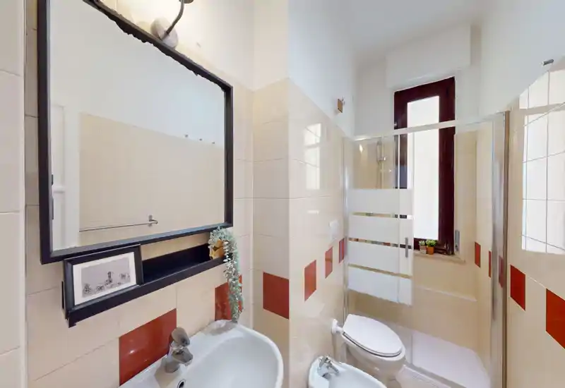 Bagno compatto e ben illuminato con lavabo, specchio, WC, bidet e cabina doccia angolare con porta in vetro; piastrelle chiare e una finestra garantiscono buona luminosità.
