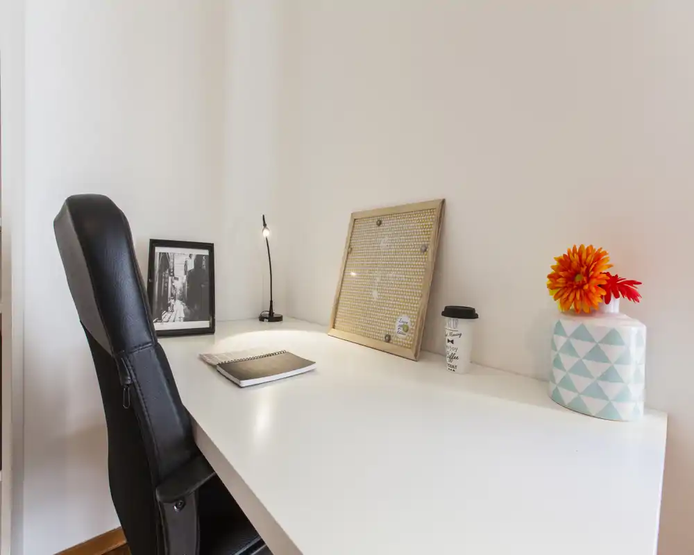 Angolo di lavoro luminoso e ordinato con scrivania bianca, comoda sedia da ufficio nera, lampada, foto incorniciata e vaso decorativo con fiori — un angolo piacevole per lavorare o studiare.