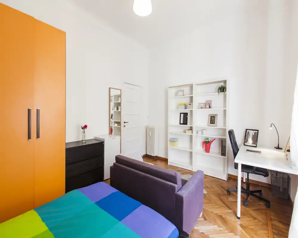 Camera da letto luminosa e ben arredata con copriletto colorato, un piccolo divano ai piedi del letto, una scrivania vicino alla finestra e scaffalature per riporre oggetti — ideale per una persona o uno studente.