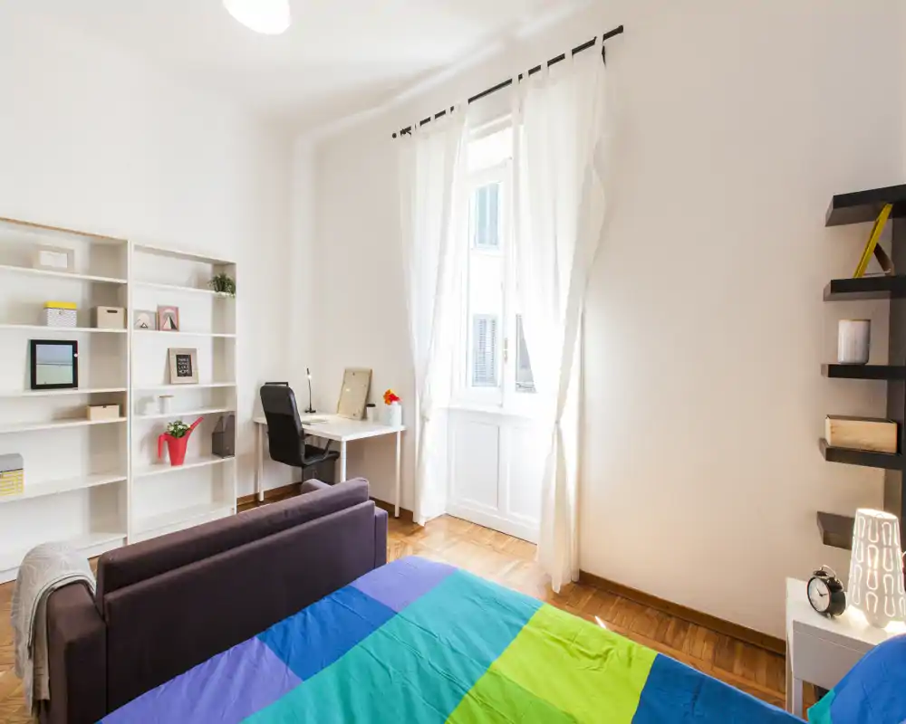 Camera da letto luminosa e moderna con copriletto colorato, piccolo divano ai piedi del letto, scaffalature e scrivania vicino a una grande finestra che dona molta luce naturale.
