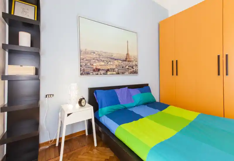 Camera da letto luminosa e colorata con letto matrimoniale, comodino e grande armadio. La stampa con la città e il pavimento in legno aggiungono carattere; perfetta per un annuncio vivace.