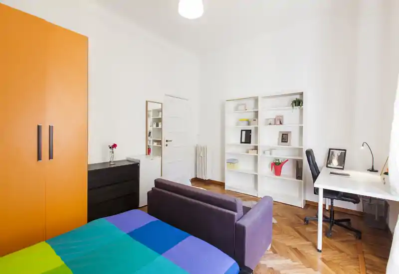 Camera da letto luminosa e ben arredata con copriletto colorato, un piccolo divano ai piedi del letto, una scrivania vicino alla finestra e scaffalature per riporre oggetti — ideale per una persona o uno studente.