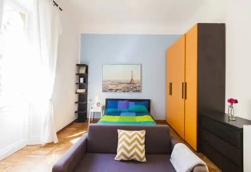 Camera da letto luminosa e colorata con letto matrimoniale al centro su una parete azzurra, un grande armadio dalle ante arancioni e luce naturale proveniente da una finestra alta che crea un ambiente caldo e moderno.