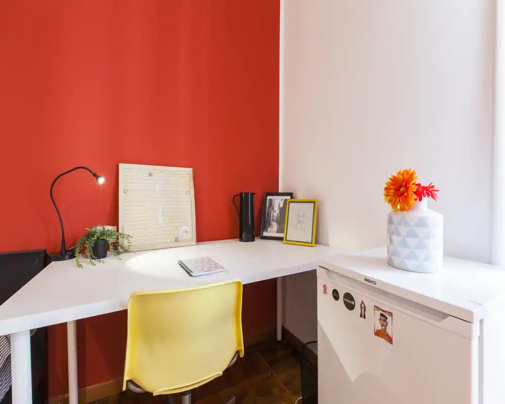Angolo studio luminoso e moderno con scrivania bianca a L, sedia gialla, lampada da tavolo e dettagli decorativi su una vivace parete rossa — perfetto per lavoro o studio.