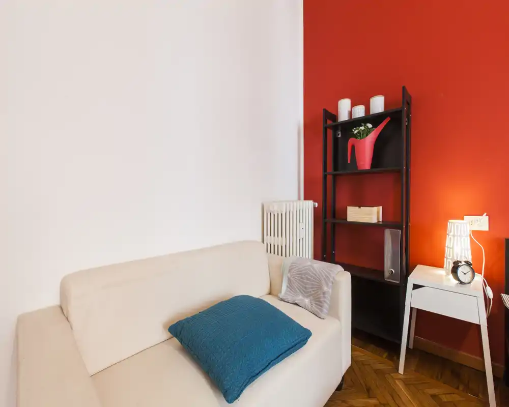 Angolo living accogliente con divano beige chiaro, cuscino turchese e una parete rossa a contrasto; presente una libreria e un tavolino con lampada, perfetto per rilassarsi.