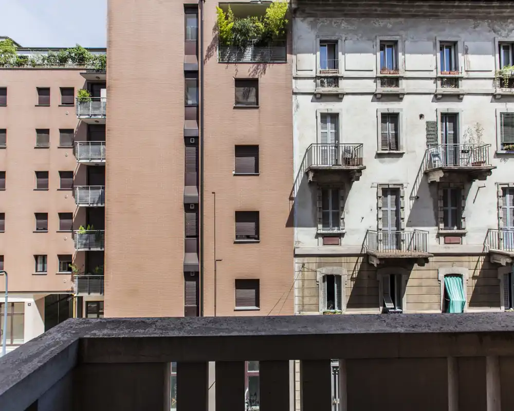 Vista da un balcone con ringhiera in cemento in primo piano e prospetti di edifici e piccoli balconi sullo sfondo.
