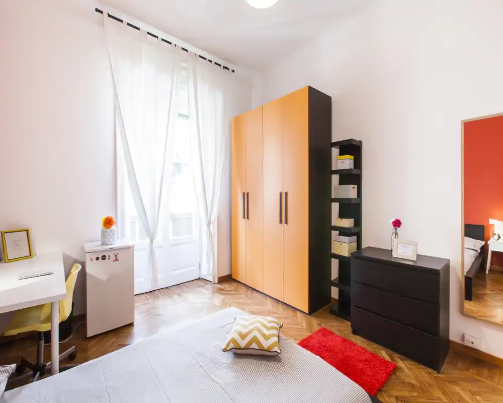 Camera da letto luminosa e ben arredata con ampia finestra, armadio arancione, comò e letto con cuscino a zigzag e tappeto rosso — perfetta per un annuncio accogliente.