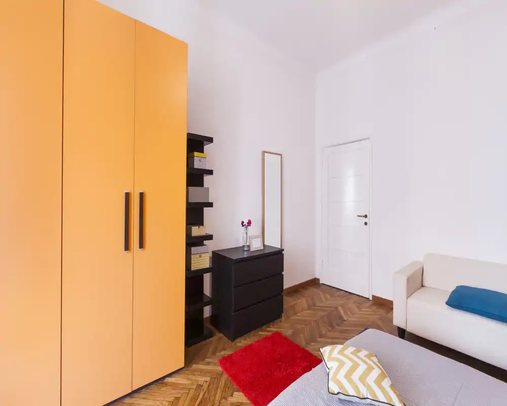 Camera da letto moderna e luminosa con letto in primo piano, armadio arancione, cassettiera e un piccolo divano. Pavimento a spina di pesce in legno e cuscini colorati con tappeto rosso che rendono l'ambiente accogliente.