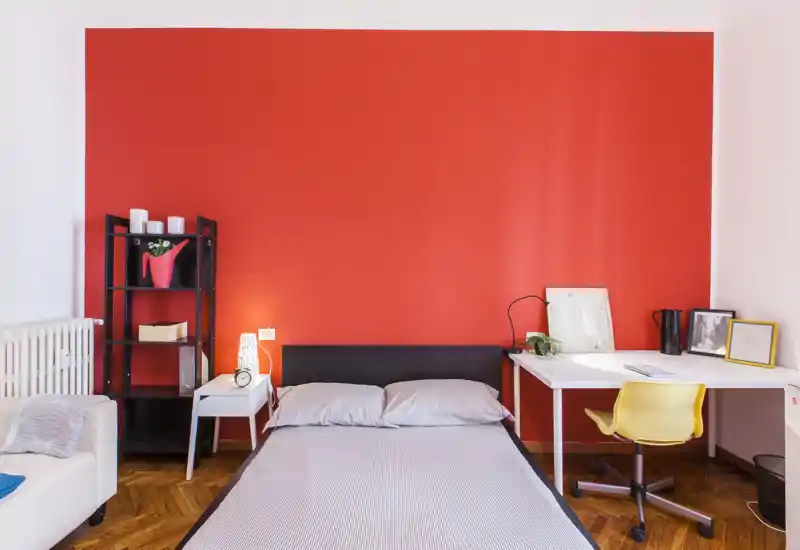 Camera da letto elegante con letto matrimoniale davanti a una parete rossa, un comodino e una scrivania con sedia gialla — luminosa, moderna e accogliente.