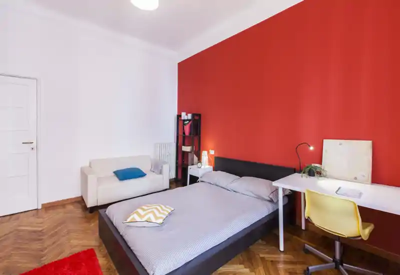 Camera da letto luminosa e moderna con letto matrimoniale davanti a una parete rossa, scrivania con sedia, piccolo divano e scaffalatura decorativa — ideale per un soggiorno confortevole.