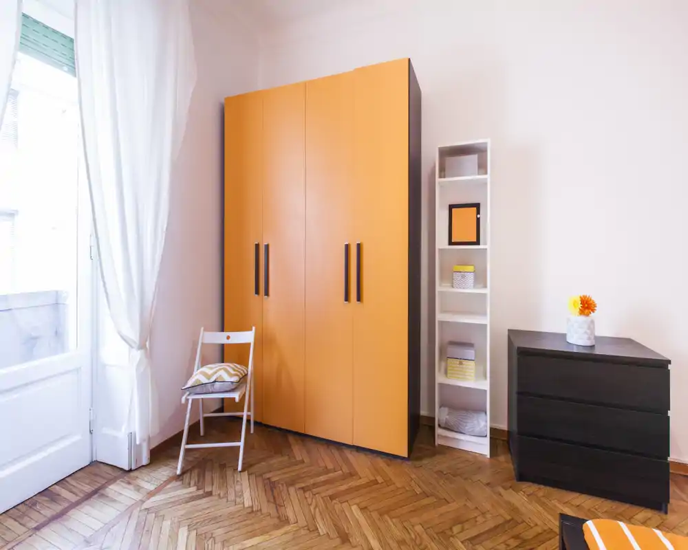 Angolo della camera luminoso e ordinato con un armadio arancione acceso, un comò e una sedia vicino alla finestra che lascia entrare luce naturale. Pavimento in parquet caldo e arredo semplice e moderno donano un ambiente accogliente.