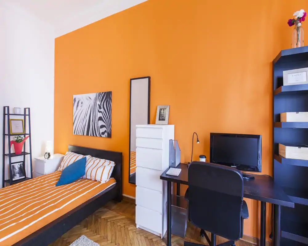 Camera da letto luminosa e stilosa con parete arancione, letto con biancheria a righe, angolo lavoro con scrivania e monitor e mensole decorative — perfetta per un annuncio accogliente.