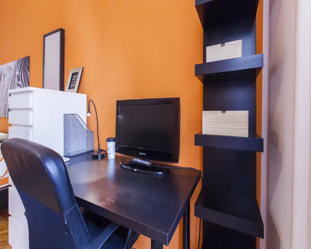 Angolo living/lavoro compatto con scrivania nera, sedia ergonomica e un monitor/TV su una calda parete arancione; scaffale e contenitori offrono pratico spazio.