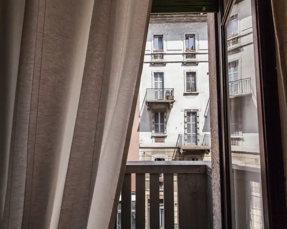 Piccolo balcone che mostra la facciata dell'edificio di fronte, inquadrata da una tenda parzialmente tirata e da una porta in vetro; luce naturale illumina la scena.