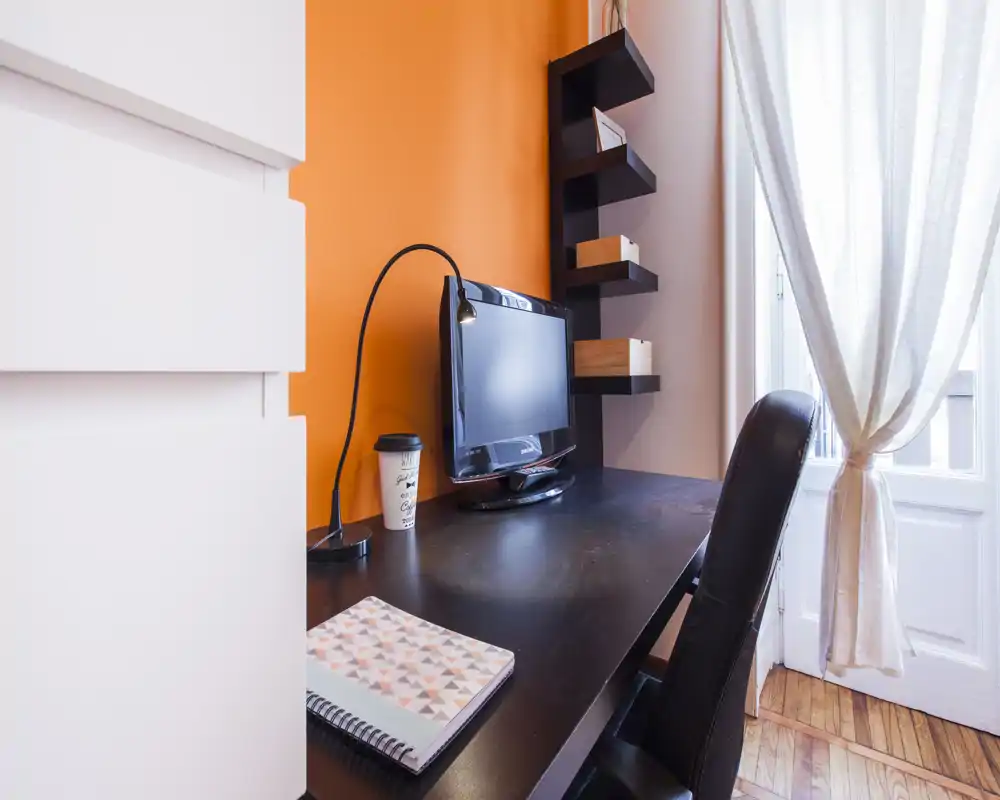 Angolo ufficio domestico compatto con scrivania nera, monitor, lampada da lavoro e sedia in pelle vicino a una finestra luminosa con tende leggere e parete arancione.