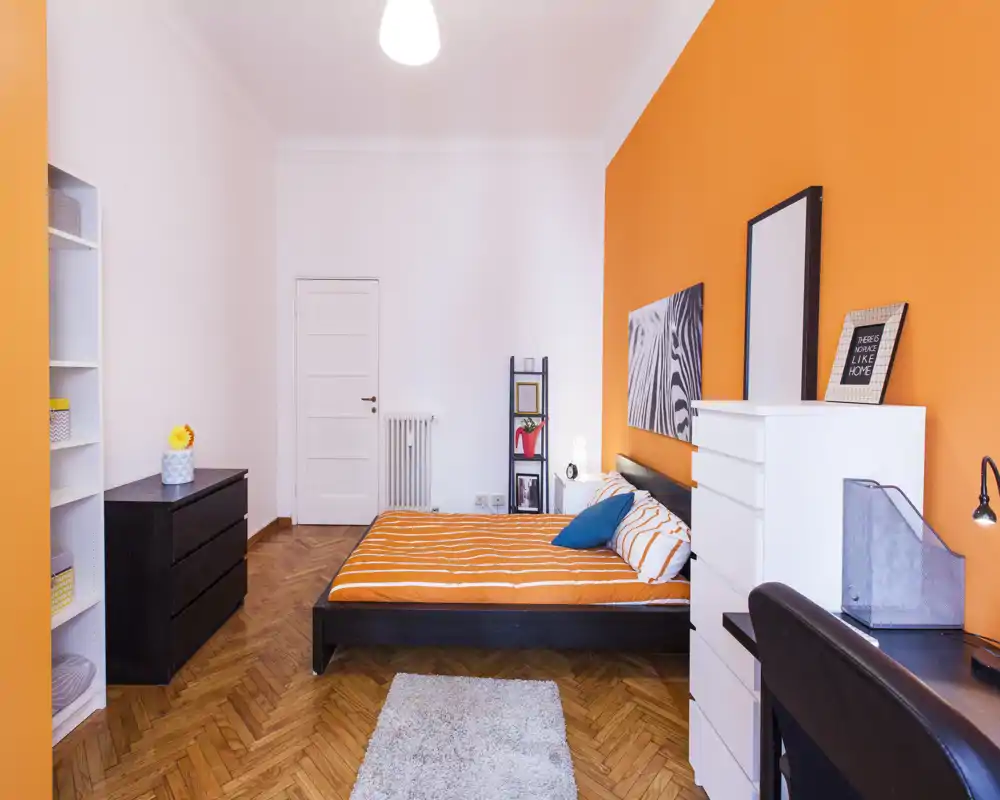 Camera da letto luminosa e moderna con parete arancione, letto matrimoniale con copriletto a righe arancioni, scaffali e scrivania — elegante e accogliente per un annuncio.