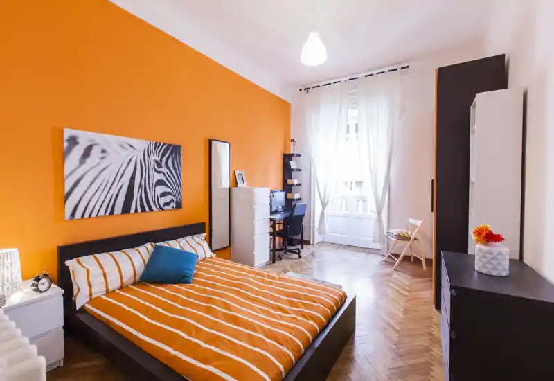 Dormitorio moderno y luminoso con pared en naranja, cama doble con ropa de cama a rayas naranjas, escritorio junto a la ventana y armarios altos — ideal para una persona o pareja que busca un espacio acogedor y bien iluminado.