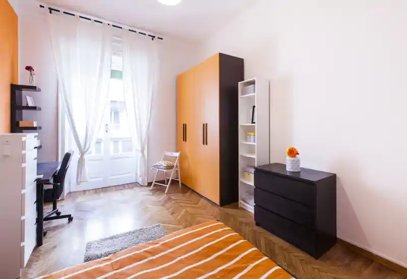 Dormitorio moderno y luminoso con acentos naranjas, cama con ropa de cama a rayas en primer plano, armario y escritorio junto a una gran ventana que aporta luz natural.