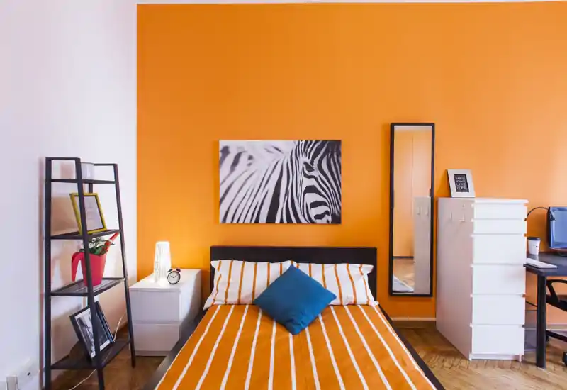 Dormitorio moderno y luminoso con pared naranja intensa, cama doble con ropa de cama a rayas y cojín azul, mesita y estantería — espacio elegante y bien iluminado, ideal para anuncios de alquiler.