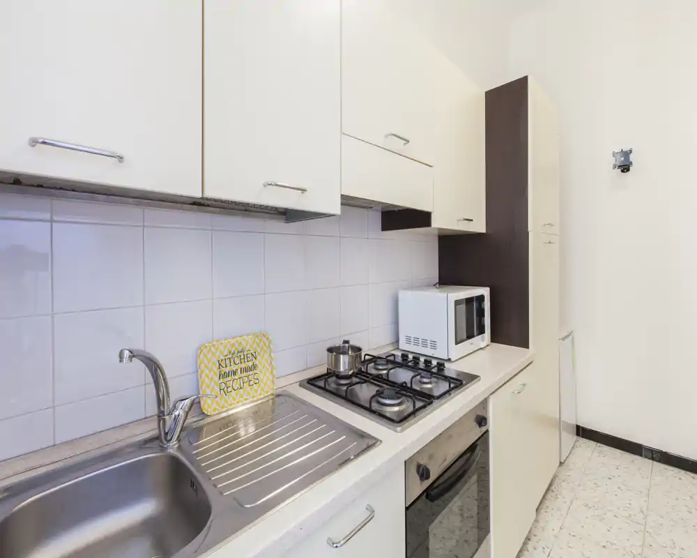 Cucina moderna e compatta con lavabo in acciaio, piano cottura a quattro fuochi, forno incassato e microonde. Pensili chiari e rivestimento a piastrelle creano uno spazio pulito e funzionale, perfetto per affitti brevi.
