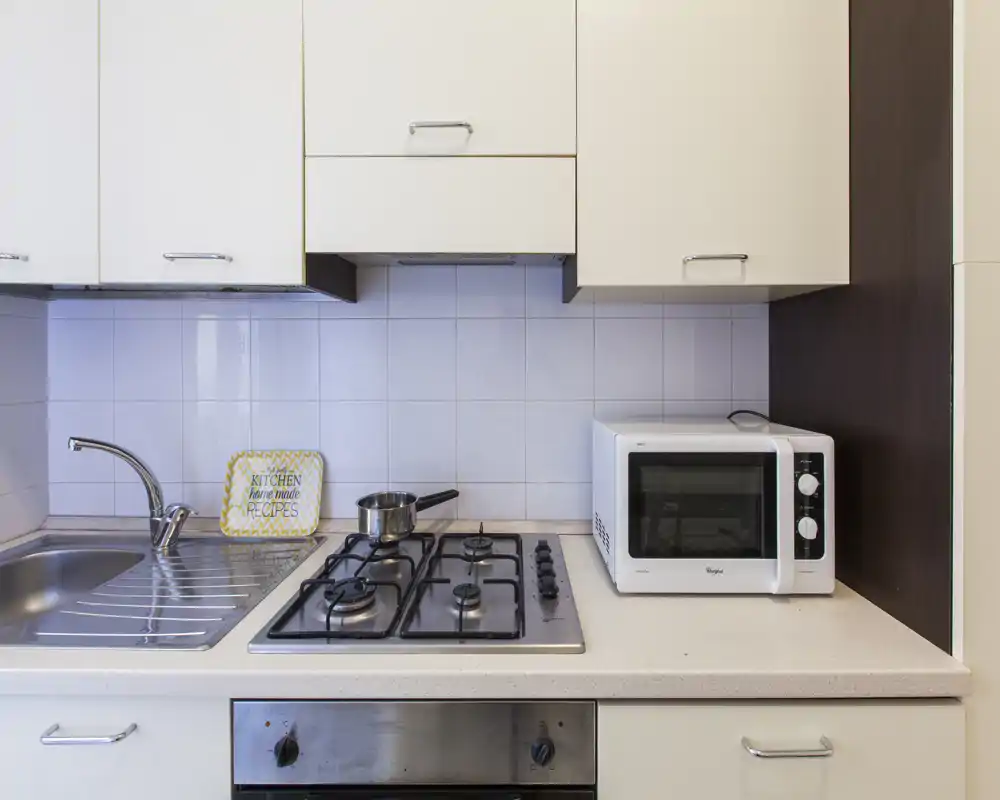 Angolo cucina compatto e moderno con lavello in acciaio, piano cottura a quattro fuochi, forno incassato e forno a microonde sul piano. Pensili neutri e piastrelle sul paraschizzi creano un’area cottura luminosa e ordinata, ideale per affitti brevi.