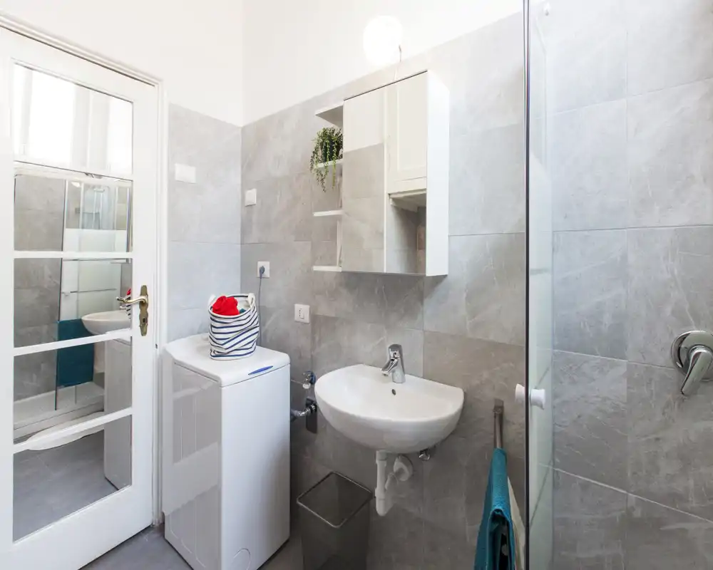 Bagno moderno e luminoso con lavabo sospeso, specchio/armadietto e una lavatrice compatta accanto. Piastrelle grigie neutre e parete doccia in vetro donano un ambiente pulito e funzionale.